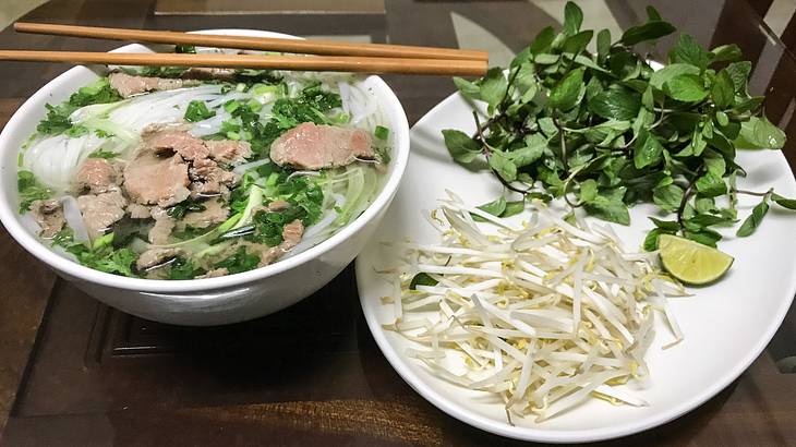 A bowl of pho noodle soup next to a plate with bean sprouts and herbs