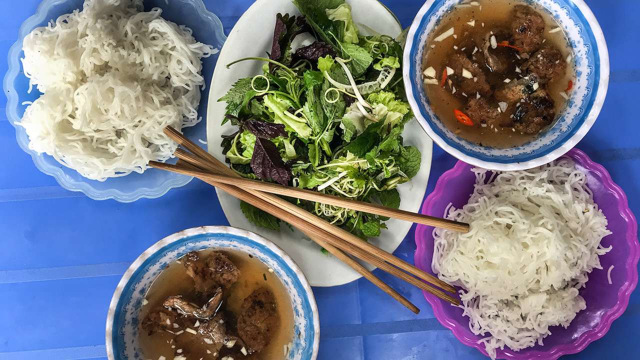 A view of a meal with noodles, meat soup, and salad on a purple cloth