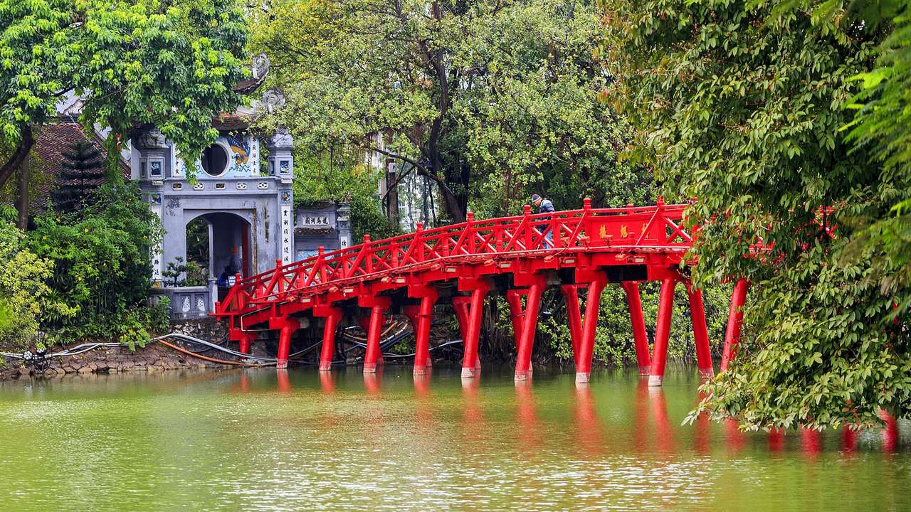 A red bridge over a lake next to a white building with greenery surrounding it