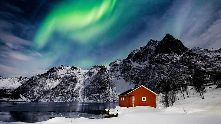 A small red cabin in the snow next to a lake and the green Northern Lights