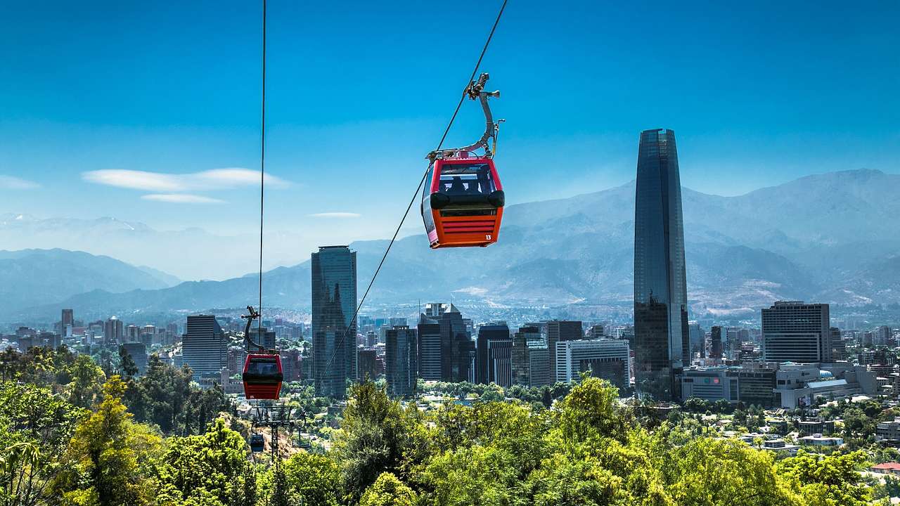 Red cable cars above greenery with city buildings and mountains behind
