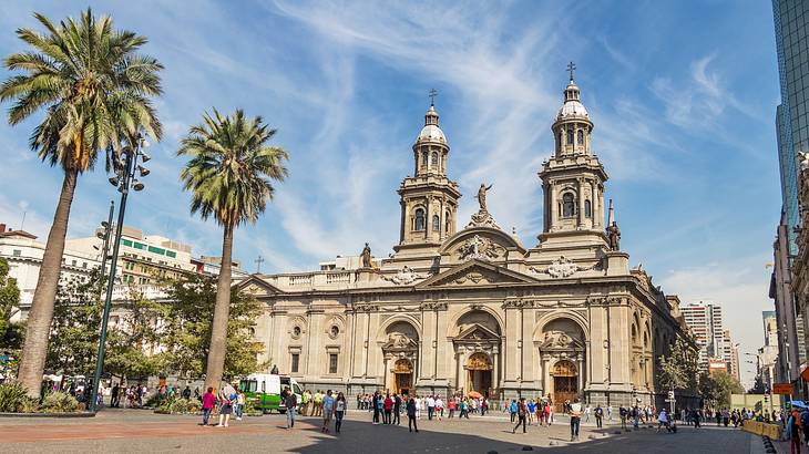 A stone cathedral with two towers, next to a square and palm trees on a nice day