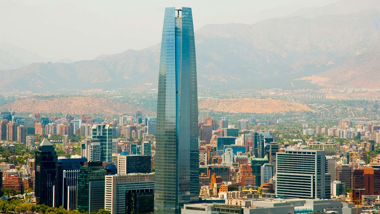 A tall mirrored skyscraper next to other smaller buildings, with mountains behind