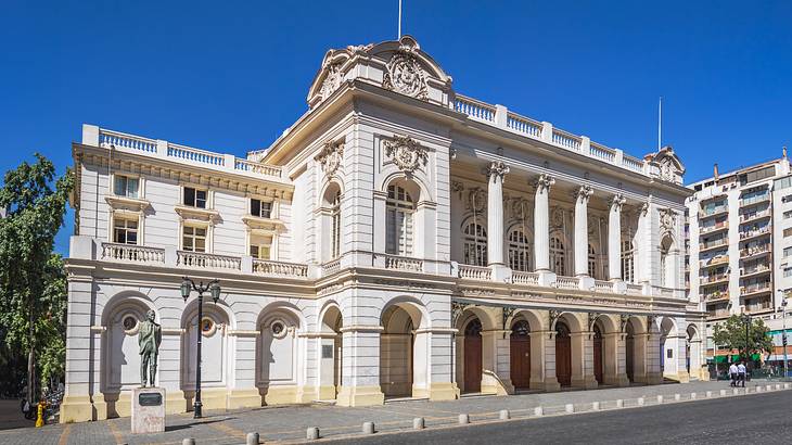 A Neoclassical stone building with arches, next to a statue and a street