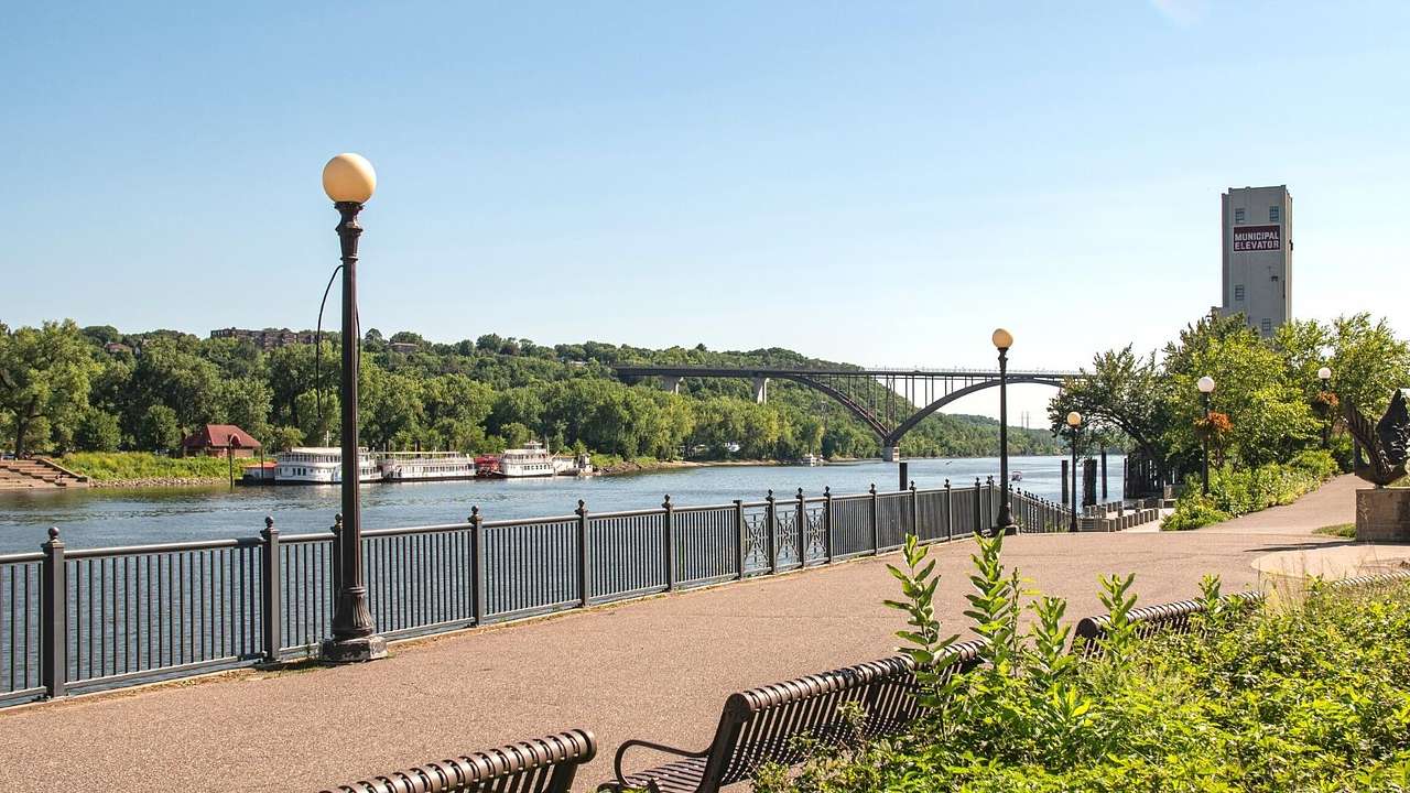 A walkway along a river with greenery surrounding it and a bridge in the distance