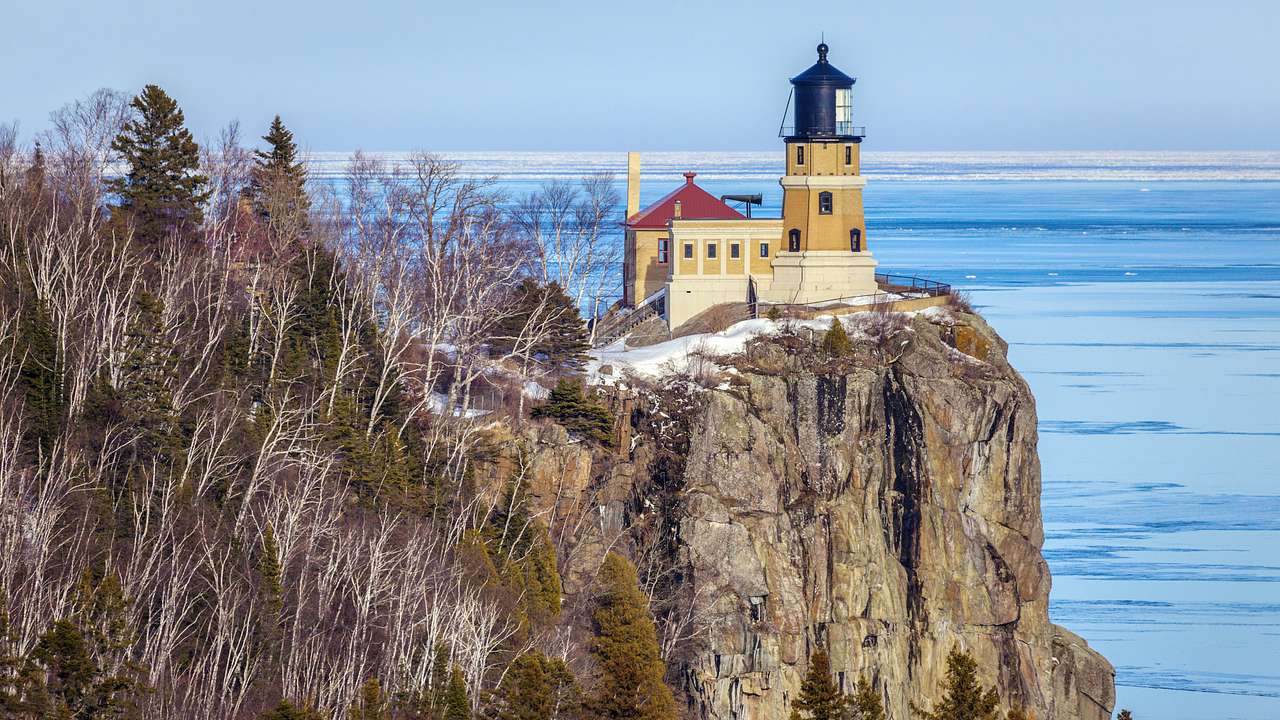 A lighthouse on a cliff with some green and bare trees next to blue water