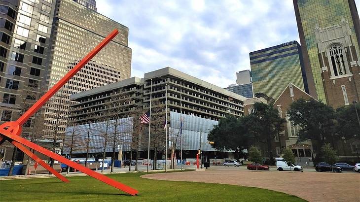 A red sculpture on the grass with city buildings behind it A red sculpture on the grass with city buildings behind it