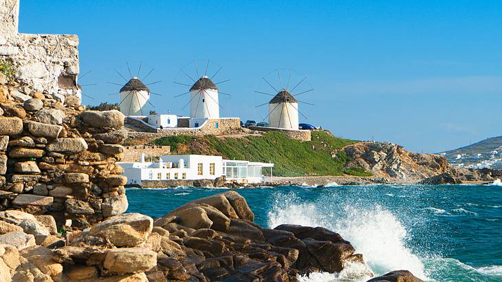 Windmills on a green hill with water and rocks in the foreground, Mykonos, Greece