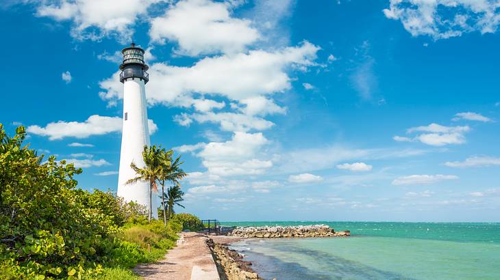 A white lighthouse on a sandy beach with greenery in front and ocean to the side