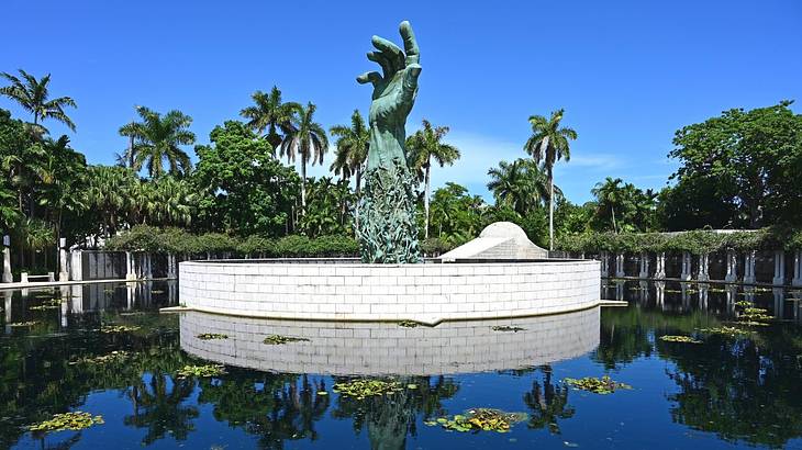 A hand sculpture in the middle of a pond with trees surrounding it