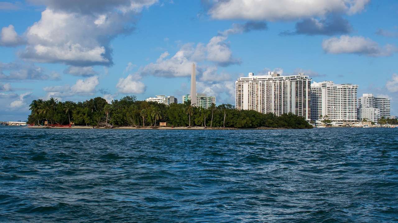 An island with trees, a tall pillar, high-rise buildings, and a partly cloudy sky