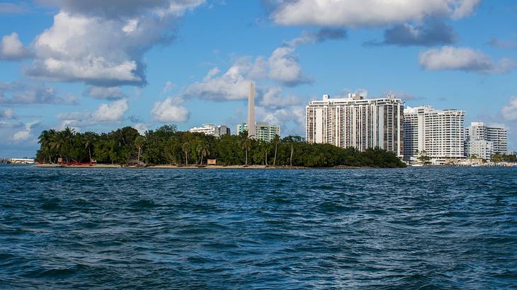 An island with trees, a tall pillar, high-rise buildings, and a partly cloudy sky