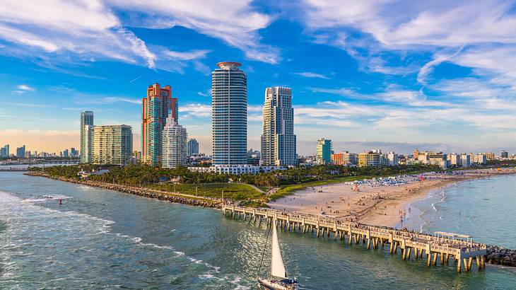 Aerial of a sailboat cruising along water with a park, beach and buildings on right