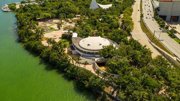 Aerial view of a park with a round roofed building, a body of water, and a road