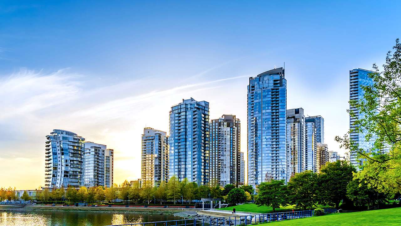 Tall glass buildings facing a body of water with a pathway and trees along it