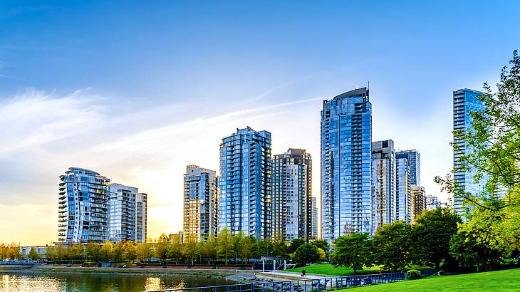 Tall glass buildings facing a body of water with a pathway and trees along it