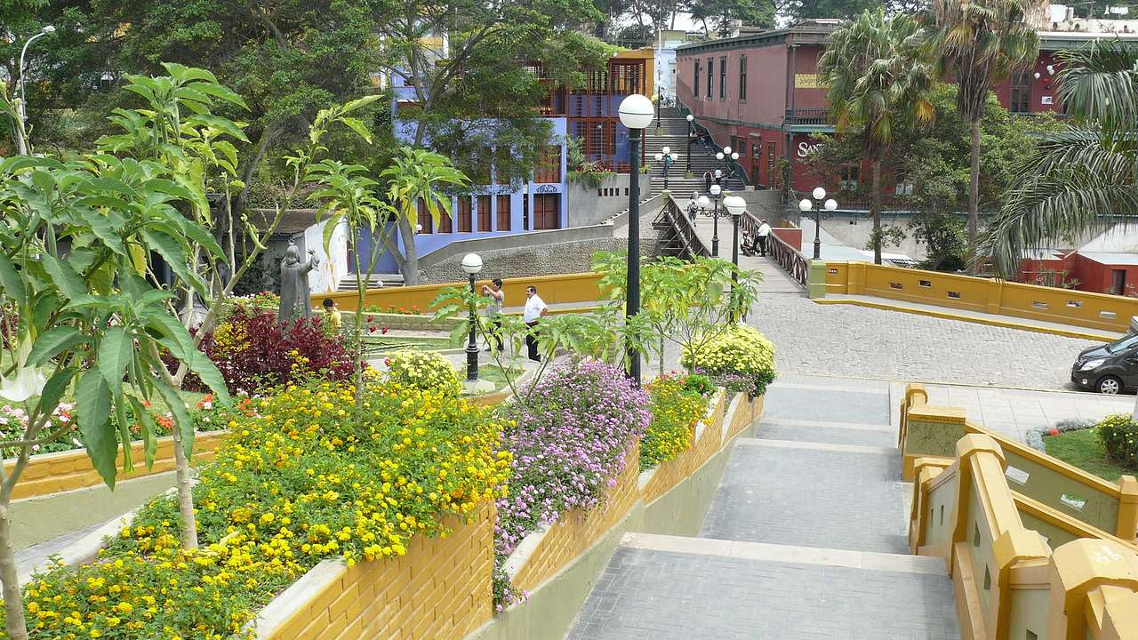 A walkway going towards a short bridge lined with streetlights, plants, and trees
