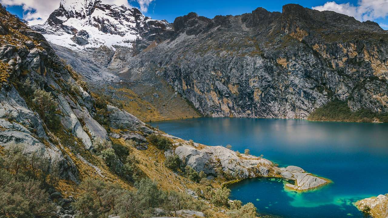 A quiet blue lake in front of a rocky mountainous landscape and a snow-capped peak