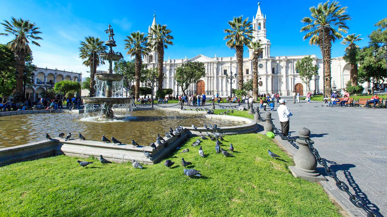 A white building with two towers facing a square with a fountain and green lawn