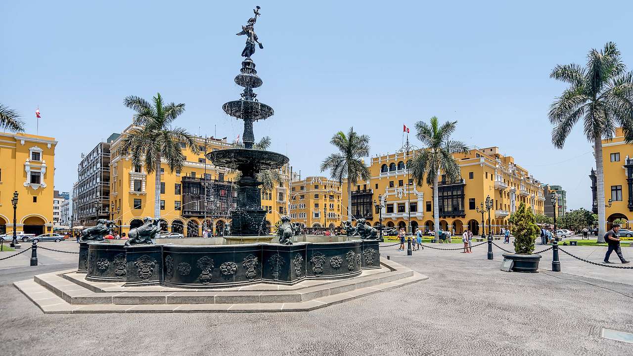 A fountain in a plaza surrounded by yellow buildings and palm trees on a sunny day