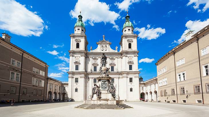 A white stone cathedral with two towers in a square under a blue sky with clouds