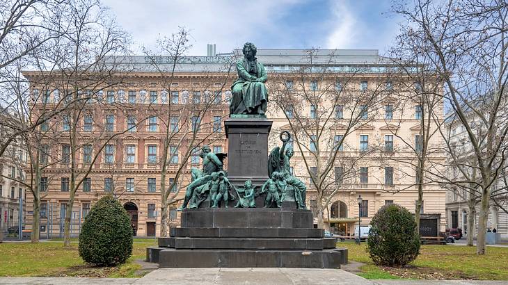 An oxidized statue of a man sitting down on a pedestal with other statues around it