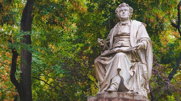 A statue of a man sitting down with a book on his lap and trees in the background