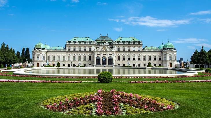 A large white palace with a green roof next to a garden with floral displays