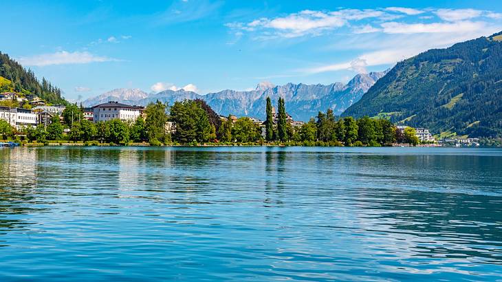 A lake next to alpine trees, greenery-covered hills, and white buildings