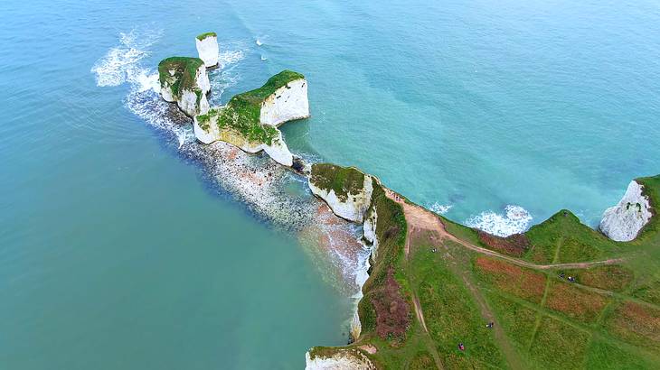 Aerial view of chalky tall cliffs topped with grass, with rocks and green water below