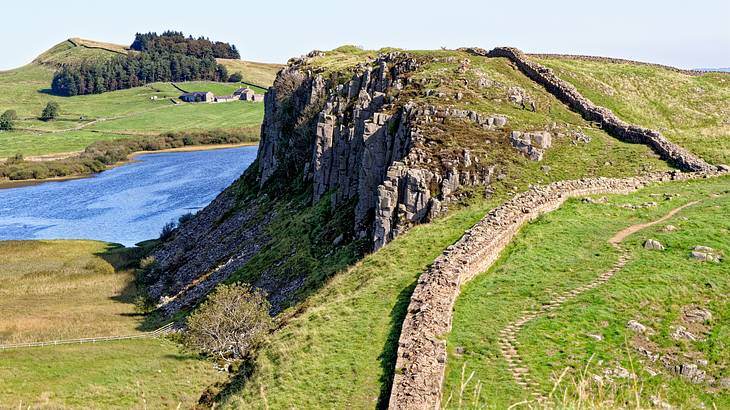 Ancient Roman wall leading up a steep, rocky green hill overlooking a body of water