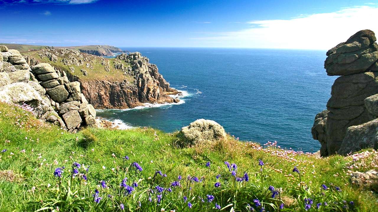 Rocky cliffs with green grass and rocks on top facing a blue sea under a blue sky