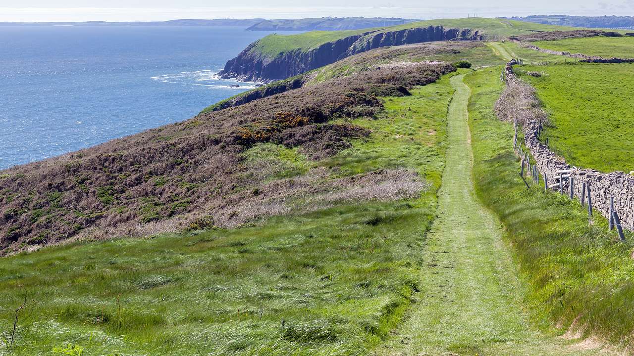 Cliffs topped by grassy fields and a path next to a blue body of water on a sunny day
