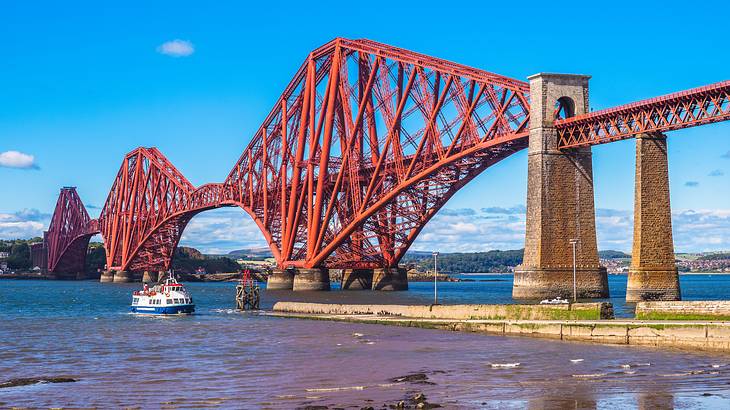 A view of a long, red, steel-structured bridge from a shore under blue sunny skies