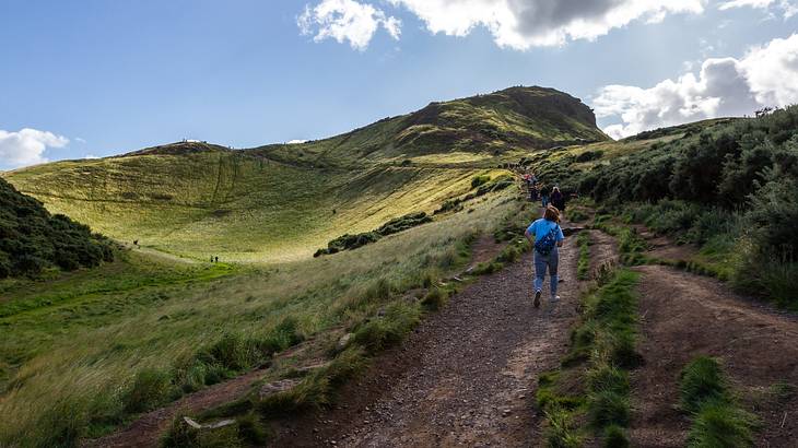 A dirt path in a hilly area leading to a mountain peak under partially cloudy skies