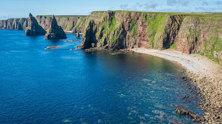 A rocky cliff partially covered in green grass next to a body of water on a sunny day