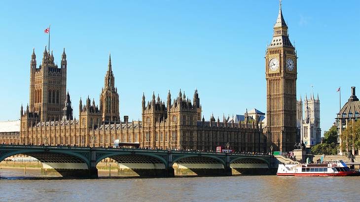 A big palace with a clock tower from across a river with a bridge and a boat