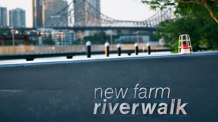 The New Farm Riverwalk sign up close with buildings and a bridge at the back