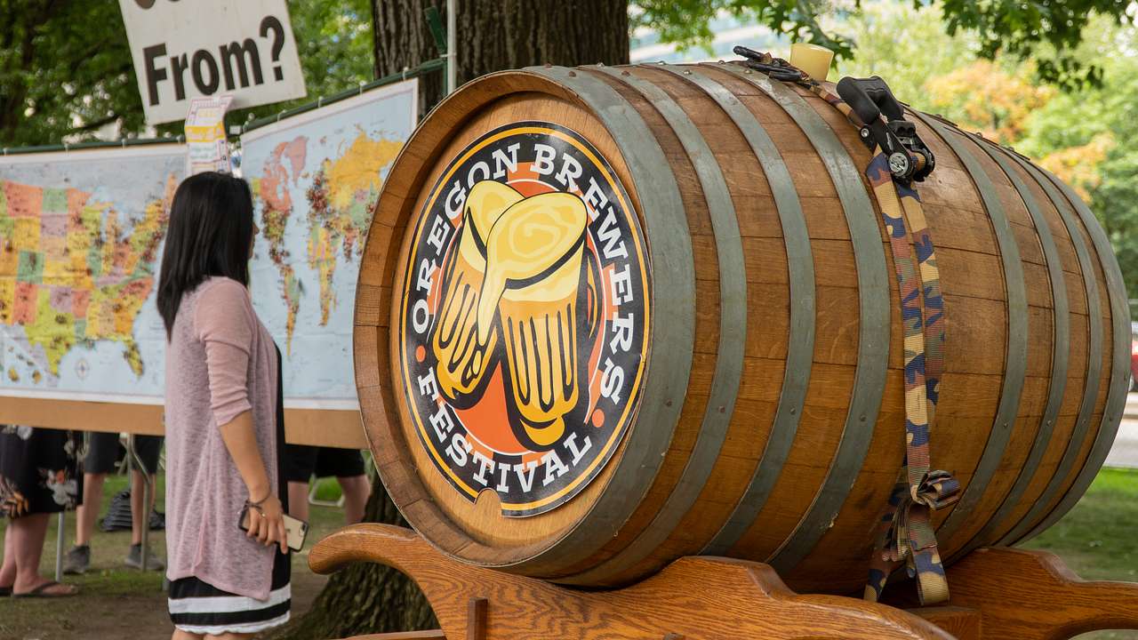 A woman standing before a map near a large wooden barrel printed with a beer logo