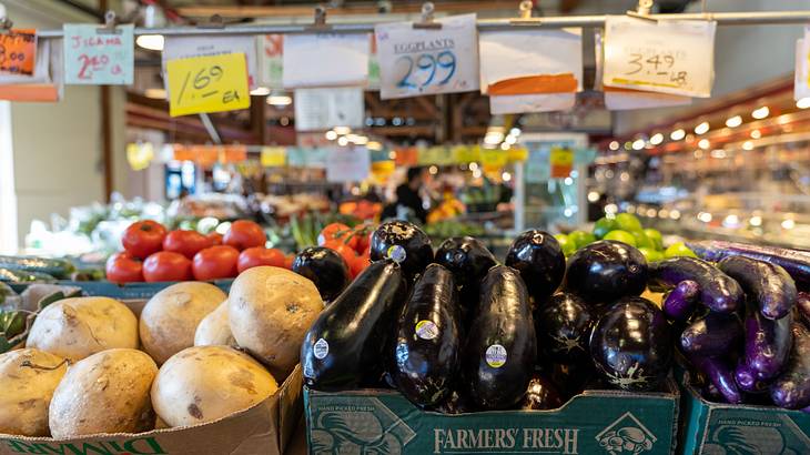 Trays of eggplants, tomatoes, and potatoes with hanging price signs in a market