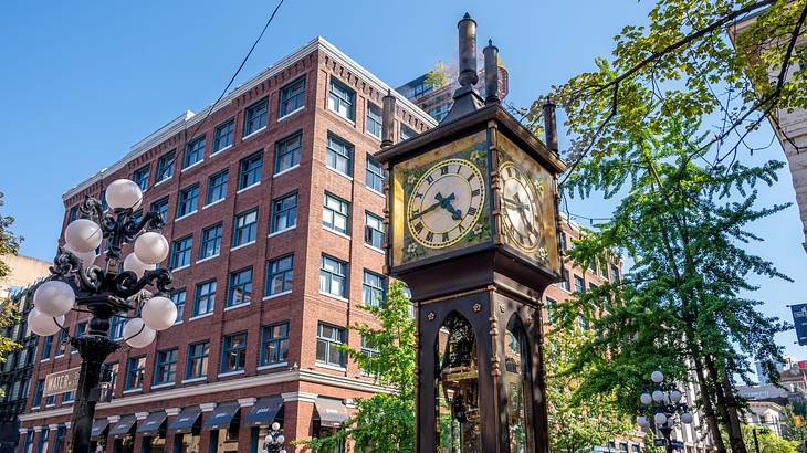 The top of a clock tower next to a street lamp, greenery, and a brick building