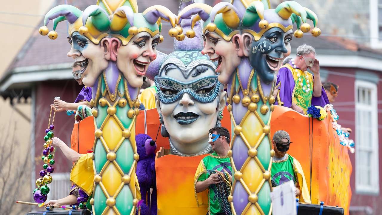 A yellow, green, and purple float with masked statues and people on it at a parade