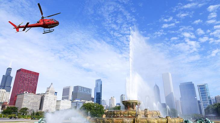 A red helicopter flying over a fountain with statues and many buildings in the back