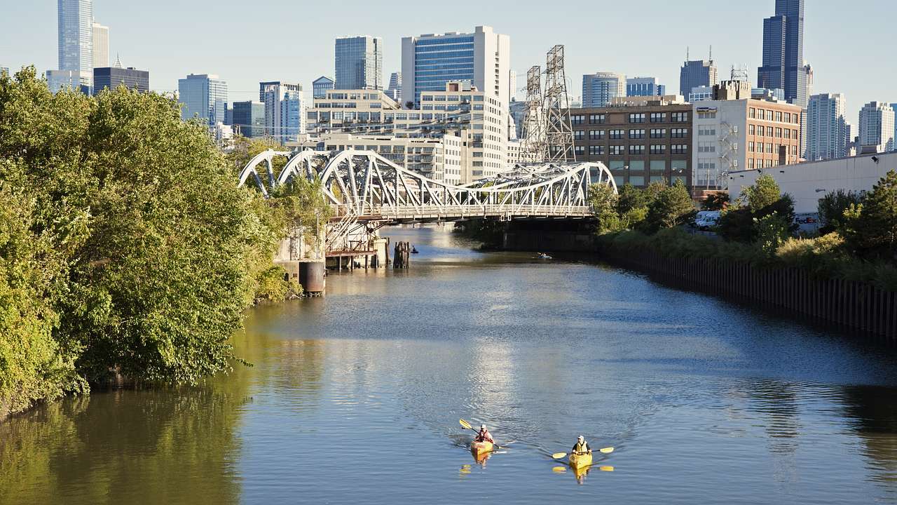 Two kayaks on a river with trees, a bridge, and an urban landscape in the background