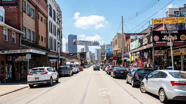 A road with cars surrounded by stores under a blue sky with some cloud