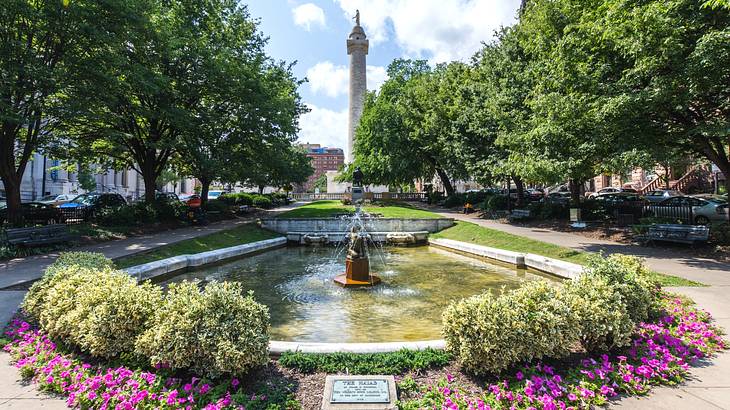 A small park with a pond and fountain, flowers, and trees, next to a stone column