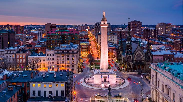 A column illuminated at night in the middle of a roundabout next to buildings