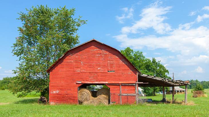 A red barn with hay near trees and grass under a blue sky with clouds