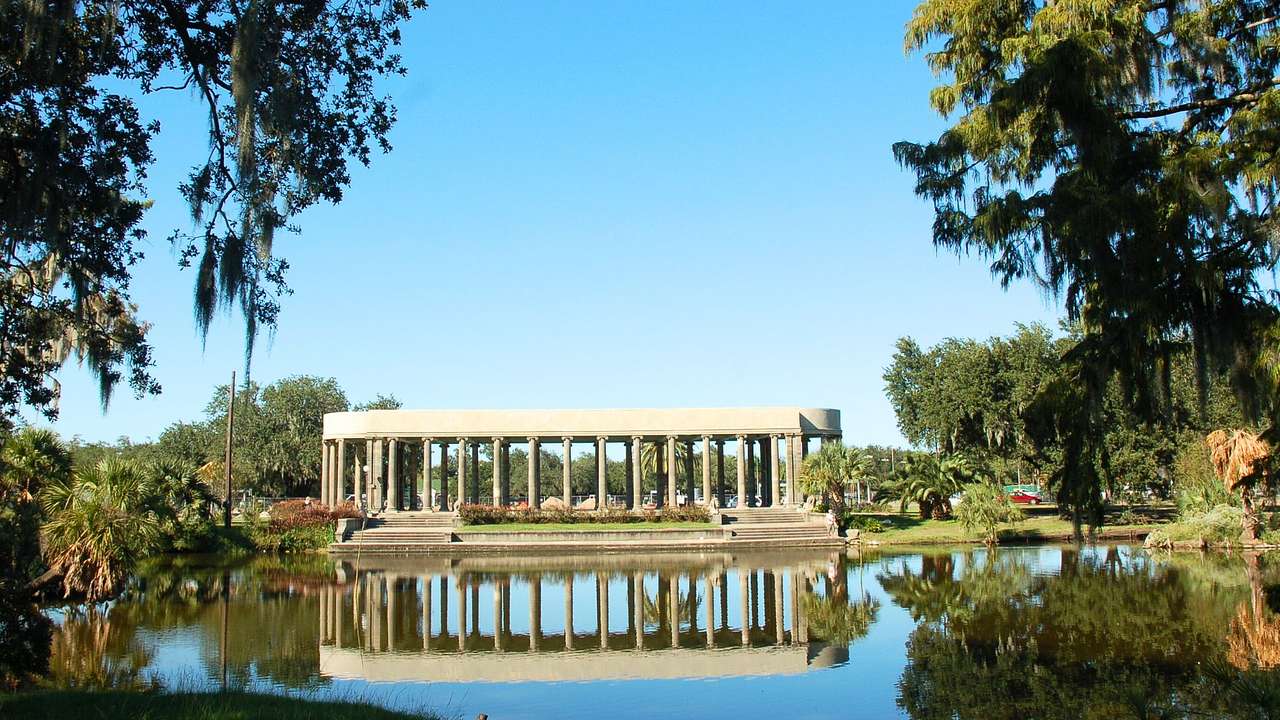 A columned open-air structure amidst a pond and trees under the clear blue sky