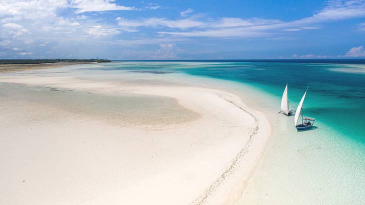 A large area of white sand next to turquoise ocean with two sailboats on it